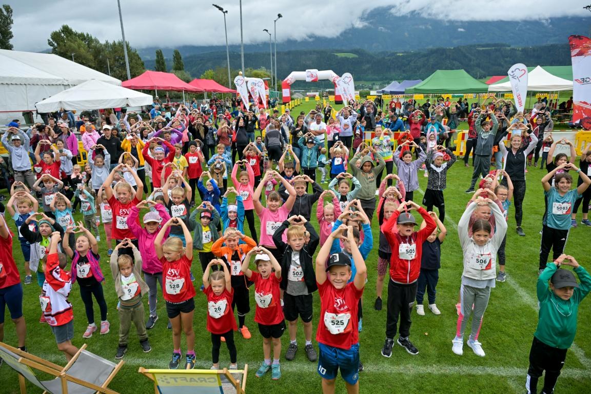 Eine Gruppe von Menschen (größtenteils Kinder) steht auf einem Sportplatz und formt mit den Händen ein Herz © C&D Moser, Foto Moser